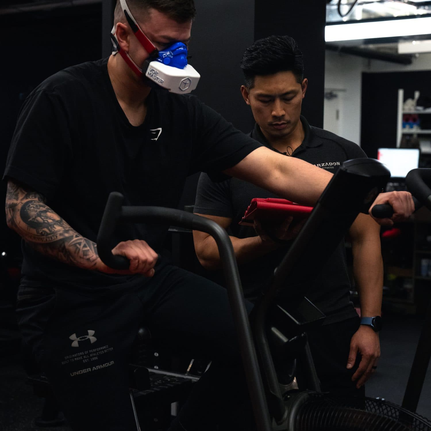 Coach monitoring a male client's VO2 max test as he pedals the air bike wearing the VO2 Master respiratory mask at Arzadon Fitness Toronto