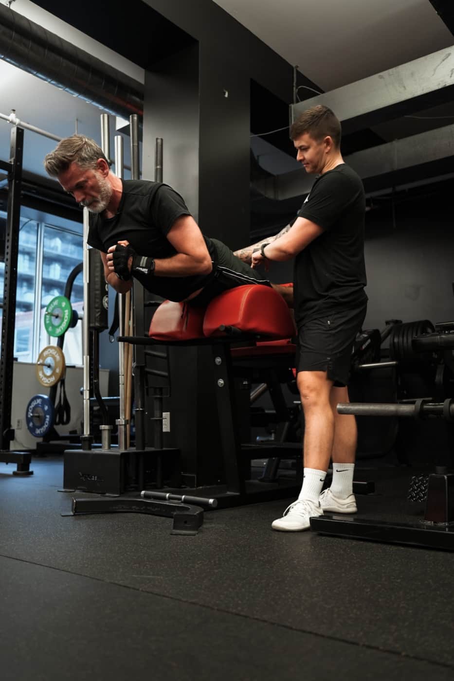 Older male client on glute-ham machine with tattooed trainer spotting