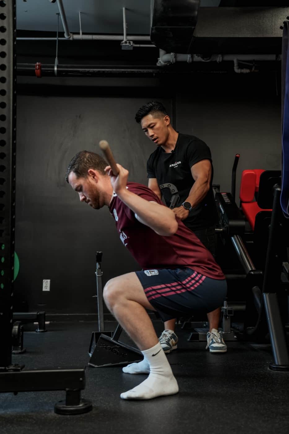 Male client swings mace in squat position while trainer coaches behind